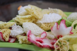 An assortment of colorful fried snacks, including various shapes and textures, predominantly in shades of yellow, white, and green, are laid out on a green surface. Some snacks have a red-pink tint, adding vibrancy to the mix.