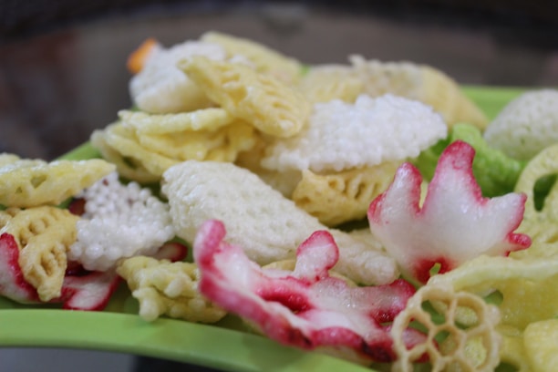 An assortment of colorful fried snacks, including various shapes and textures, predominantly in shades of yellow, white, and green, are laid out on a green surface. Some snacks have a red-pink tint, adding vibrancy to the mix.