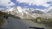 A scenic mountain landscape features snow-capped peaks under a partly cloudy sky. A winding road runs through the rugged terrain, with two people standing beside a parked motorcycle on the roadside. A sign on the right reads 'Bara-lacha la Pass Altitude 4650 M'.