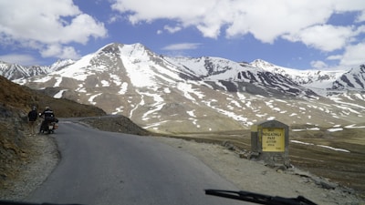 A scenic mountain landscape features snow-capped peaks under a partly cloudy sky. A winding road runs through the rugged terrain, with two people standing beside a parked motorcycle on the roadside. A sign on the right reads 'Bara-lacha la Pass Altitude 4650 M'.