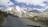 A scenic mountain landscape features snow-capped peaks under a partly cloudy sky. A winding road runs through the rugged terrain, with two people standing beside a parked motorcycle on the roadside. A sign on the right reads 'Bara-lacha la Pass Altitude 4650 M'.