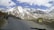 A scenic mountain landscape features snow-capped peaks under a partly cloudy sky. A winding road runs through the rugged terrain, with two people standing beside a parked motorcycle on the roadside. A sign on the right reads 'Bara-lacha la Pass Altitude 4650 M'.