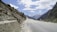 A rocky mountain road stretches into the distance with a backdrop of snow-capped peaks. The sky is filled with fluffy white clouds contrasting against the blue. Sparse greenery is visible along the road, with large rocky slopes on one side.