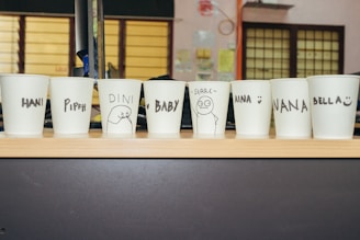 Several white paper cups placed in a row on a wooden surface, each with a name or drawing in black marker. The names include Hani, Pipah, Dini, Baby, Aina, Nana, and Bella. Some have simple illustrations next to the names.