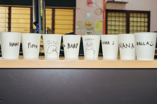Several white paper cups placed in a row on a wooden surface, each with a name or drawing in black marker. The names include Hani, Pipah, Dini, Baby, Aina, Nana, and Bella. Some have simple illustrations next to the names.