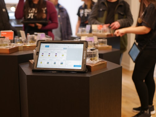 A tablet displaying a menu is positioned on a dark pedestal in a store setting. In the background, various jars with products are arranged on similar pedestals, and several people are interacting with the items, dressed casually.