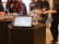 A tablet displaying a menu is positioned on a dark pedestal in a store setting. In the background, various jars with products are arranged on similar pedestals, and several people are interacting with the items, dressed casually.