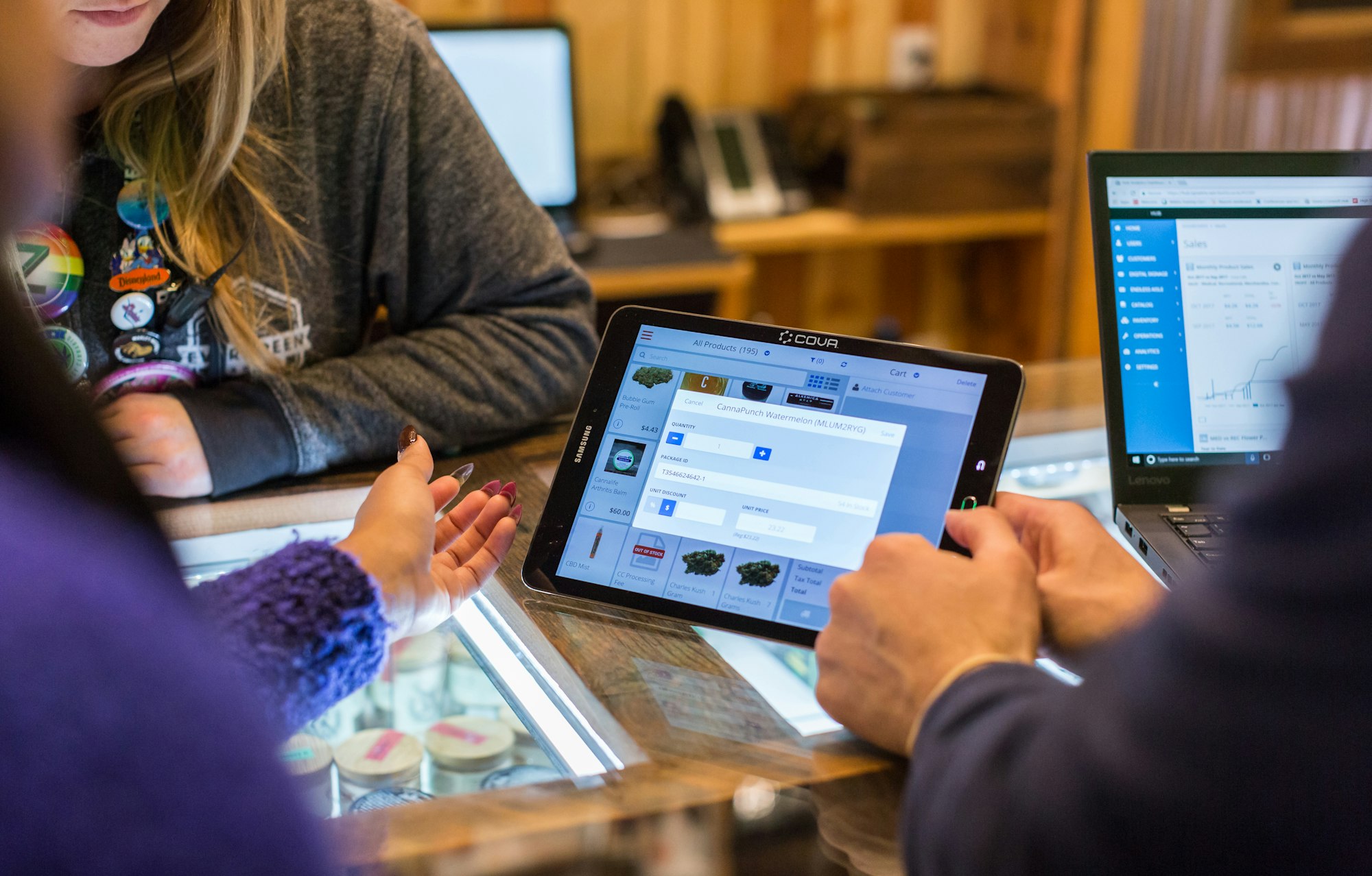 Two cannabis customers discuss product options with a female budtender across a dispensary sales counter. Between them is a lit tablet displaying Cova's point of sale cannabis software.