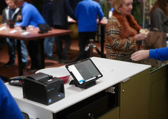A modern retail checkout setup featuring a digital tablet on a stand displaying information, alongside a card payment terminal and a receipt printer. In the background, several people interact in a busy store environment, with some blurred movement suggesting activity.