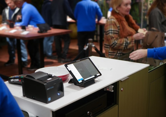 A modern retail checkout setup featuring a digital tablet on a stand displaying information, alongside a card payment terminal and a receipt printer. In the background, several people interact in a busy store environment, with some blurred movement suggesting activity.