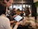 Two people are interacting in a retail setting, possibly a store. One is holding a tablet, displaying information. Various products in glass cases are visible in the foreground, and other people are in the background.