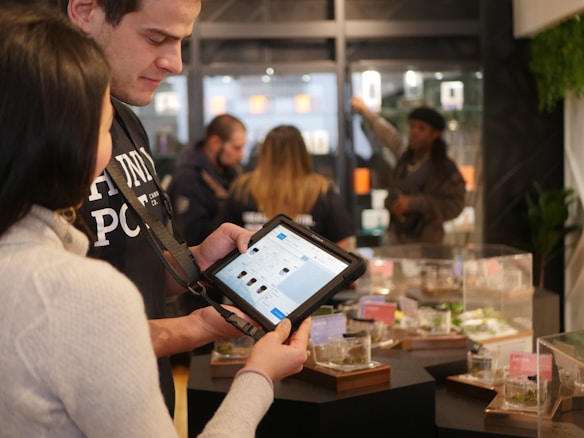 Two people are interacting in a retail setting, possibly a store. One is holding a tablet, displaying information. Various products in glass cases are visible in the foreground, and other people are in the background.
