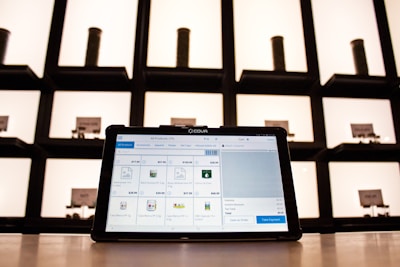 A tablet displaying a point-of-sale application sits on a countertop, with shelves in the background. The shelves are illuminated and contain cylindrical containers.
