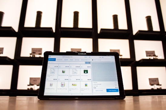 A tablet displaying a point-of-sale application sits on a countertop, with shelves in the background. The shelves are illuminated and contain cylindrical containers.