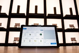 A tablet displaying a point-of-sale application sits on a countertop, with shelves in the background. The shelves are illuminated and contain cylindrical containers.