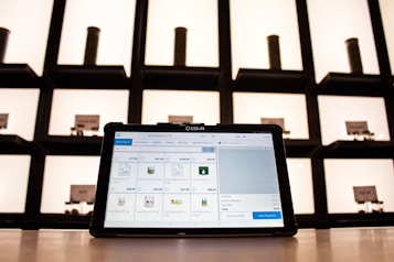 A tablet displaying a point-of-sale application sits on a countertop, with shelves in the background. The shelves are illuminated and contain cylindrical containers.