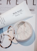 A close-up scene featuring a tube labeled 'Holy Grail Beauty Co.' and a tub of cream on top of a magazine or printed material. There is also a pair of tortoiseshell hoop earrings partially in view. The cream appears to be a facial or skincare product, with a smooth and creamy texture.