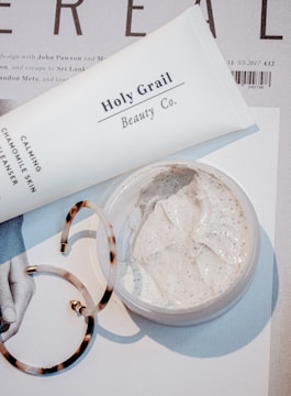 A close-up scene featuring a tube labeled 'Holy Grail Beauty Co.' and a tub of cream on top of a magazine or printed material. There is also a pair of tortoiseshell hoop earrings partially in view. The cream appears to be a facial or skincare product, with a smooth and creamy texture.