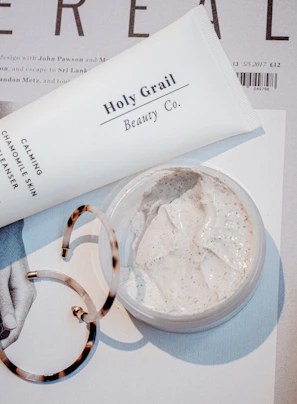 A close-up scene featuring a tube labeled 'Holy Grail Beauty Co.' and a tub of cream on top of a magazine or printed material. There is also a pair of tortoiseshell hoop earrings partially in view. The cream appears to be a facial or skincare product, with a smooth and creamy texture.