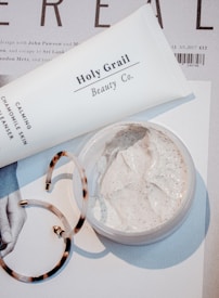 A close-up scene featuring a tube labeled 'Holy Grail Beauty Co.' and a tub of cream on top of a magazine or printed material. There is also a pair of tortoiseshell hoop earrings partially in view. The cream appears to be a facial or skincare product, with a smooth and creamy texture.