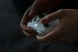 Hands are holding a white gaming controller with illuminated buttons in a dimly lit environment. The fingers are poised to press the buttons, suggesting engagement in a gaming activity.