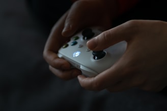 Hands are holding a white gaming controller with illuminated buttons in a dimly lit environment. The fingers are poised to press the buttons, suggesting engagement in a gaming activity.