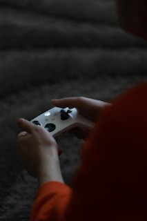 A person holding a white gaming controller with both hands, set against a dimly lit room with a textured carpet. The figure is partially in shadow, adding a sense of focus on the controller and hands.