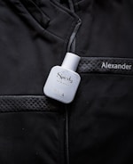 A white bottle labeled 'Sporty' eau de parfum is placed on a black jacket with quilted details. The name 'Alexander' is embroidered or printed on the jacket.
