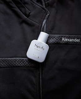 A white bottle labeled 'Sporty' eau de parfum is placed on a black jacket with quilted details. The name 'Alexander' is embroidered or printed on the jacket.