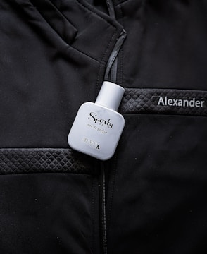A white bottle labeled 'Sporty' eau de parfum is placed on a black jacket with quilted details. The name 'Alexander' is embroidered or printed on the jacket.