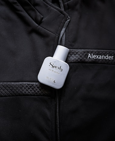 A white bottle labeled 'Sporty' eau de parfum is placed on a black jacket with quilted details. The name 'Alexander' is embroidered or printed on the jacket.