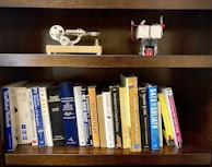 A wooden bookshelf holding a collection of books on various topics such as programming, dictionaries, and computer science. On the top shelf, there are two small mechanical models, one resembling a stirling engine and the other appearing to be a small engine with a propeller.