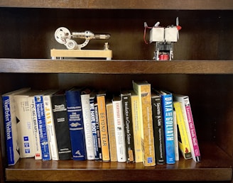 A wooden bookshelf holding a collection of books on various topics such as programming, dictionaries, and computer science. On the top shelf, there are two small mechanical models, one resembling a stirling engine and the other appearing to be a small engine with a propeller.