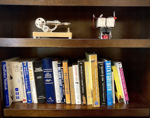 A wooden bookshelf holding a collection of books on various topics such as programming, dictionaries, and computer science. On the top shelf, there are two small mechanical models, one resembling a stirling engine and the other appearing to be a small engine with a propeller.