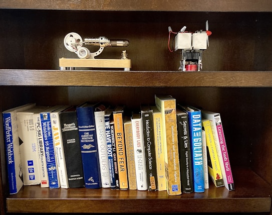 A wooden bookshelf holding a collection of books on various topics such as programming, dictionaries, and computer science. On the top shelf, there are two small mechanical models, one resembling a stirling engine and the other appearing to be a small engine with a propeller.