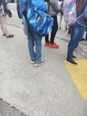 A group of people, likely students, stand on a sidewalk. They are dressed casually with backpacks. The ground is concrete with a yellow tactile paving strip. Some characteristically colorful shoes and backpacks are visible.