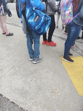 A group of people, likely students, stand on a sidewalk. They are dressed casually with backpacks. The ground is concrete with a yellow tactile paving strip. Some characteristically colorful shoes and backpacks are visible.