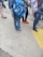 A group of people, likely students, stand on a sidewalk. They are dressed casually with backpacks. The ground is concrete with a yellow tactile paving strip. Some characteristically colorful shoes and backpacks are visible.