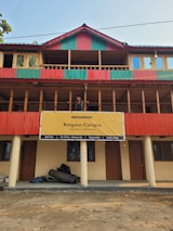 A multi-story building with a prominent red and green striped design on its exterior. A large yellow banner hangs on the building with the text 'INDIAHIKES Kotgaon Campus 6,455 feet | 1,967 meters'. Below, rolled-up materials rest against the building's wall. A man stands on the balcony, looking out at the surroundings.