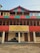 A multi-story building with a prominent red and green striped design on its exterior. A large yellow banner hangs on the building with the text 'INDIAHIKES Kotgaon Campus 6,455 feet | 1,967 meters'. Below, rolled-up materials rest against the building's wall. A man stands on the balcony, looking out at the surroundings.