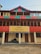 A multi-story building with a prominent red and green striped design on its exterior. A large yellow banner hangs on the building with the text 'INDIAHIKES Kotgaon Campus 6,455 feet | 1,967 meters'. Below, rolled-up materials rest against the building's wall. A man stands on the balcony, looking out at the surroundings.