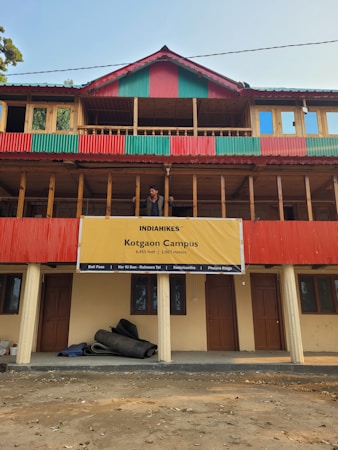A multi-story building with a prominent red and green striped design on its exterior. A large yellow banner hangs on the building with the text 'INDIAHIKES Kotgaon Campus 6,455 feet | 1,967 meters'. Below, rolled-up materials rest against the building's wall. A man stands on the balcony, looking out at the surroundings.