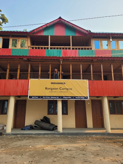 A multi-story building with a prominent red and green striped design on its exterior. A large yellow banner hangs on the building with the text 'INDIAHIKES Kotgaon Campus 6,455 feet | 1,967 meters'. Below, rolled-up materials rest against the building's wall. A man stands on the balcony, looking out at the surroundings.