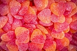 A close-up view of a pile of heart-shaped jelly candies coated in sugar. The candies are colored in vibrant shades of red and orange, creating a visually appealing texture.