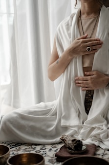 A person is seated in a meditative posture with hands placed on the chest and abdomen. The individual is draped in white, flowing fabric and is adorned with a ring and necklace. A soft, natural light filters through the curtains in the background. In the foreground, there are decorative bowls and what appears to be a piece of smudge stick resting on a small plate.