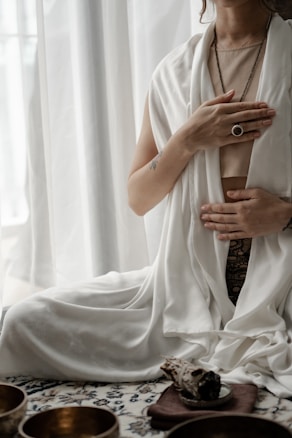 A person is seated in a meditative posture with hands placed on the chest and abdomen. The individual is draped in white, flowing fabric and is adorned with a ring and necklace. A soft, natural light filters through the curtains in the background. In the foreground, there are decorative bowls and what appears to be a piece of smudge stick resting on a small plate.