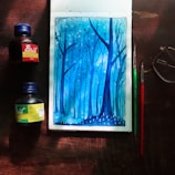 A watercolor painting depicting a serene forest scene in shades of blue, with tall, slender trees and small dots that resemble falling leaves or snow. The artwork is surrounded by art supplies including two bottles of ink, one red and one green, and two paintbrushes. A pair of glasses is placed nearby, all on a wooden surface.
