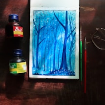 A watercolor painting depicting a serene forest scene in shades of blue, with tall, slender trees and small dots that resemble falling leaves or snow. The artwork is surrounded by art supplies including two bottles of ink, one red and one green, and two paintbrushes. A pair of glasses is placed nearby, all on a wooden surface.
