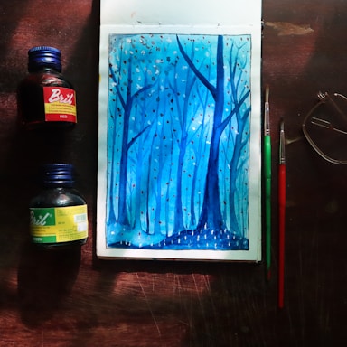 A watercolor painting depicting a serene forest scene in shades of blue, with tall, slender trees and small dots that resemble falling leaves or snow. The artwork is surrounded by art supplies including two bottles of ink, one red and one green, and two paintbrushes. A pair of glasses is placed nearby, all on a wooden surface.