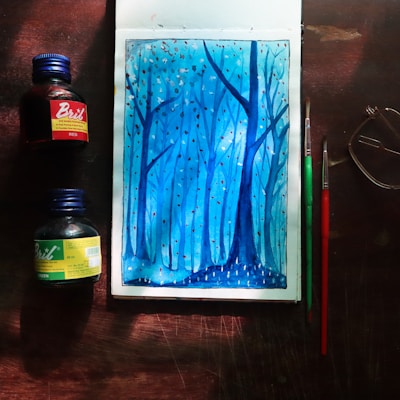 A watercolor painting depicting a serene forest scene in shades of blue, with tall, slender trees and small dots that resemble falling leaves or snow. The artwork is surrounded by art supplies including two bottles of ink, one red and one green, and two paintbrushes. A pair of glasses is placed nearby, all on a wooden surface.